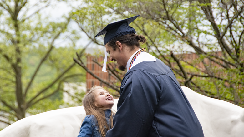 A man in academic regalia including picks up his small daughter in front of Schuylkill's lion shrine.